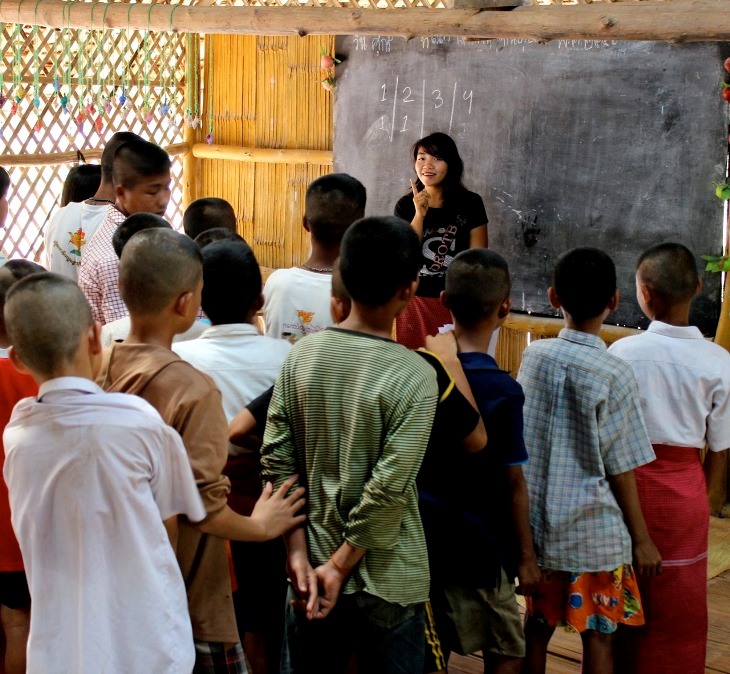 A young female student teaches in front of a class of boys, with a blackboard in the background.